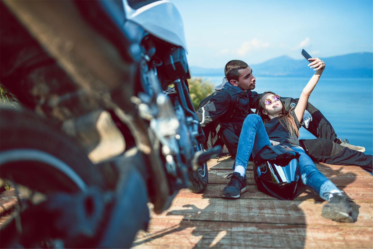 Two young people taking a selfie by a cliff near their motorcycle.